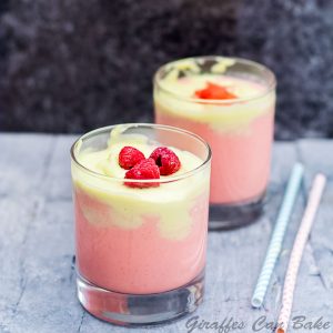 Short glass of smoothie with pink and green layer, topped with raspberries. Another short glass in background - square cropped
